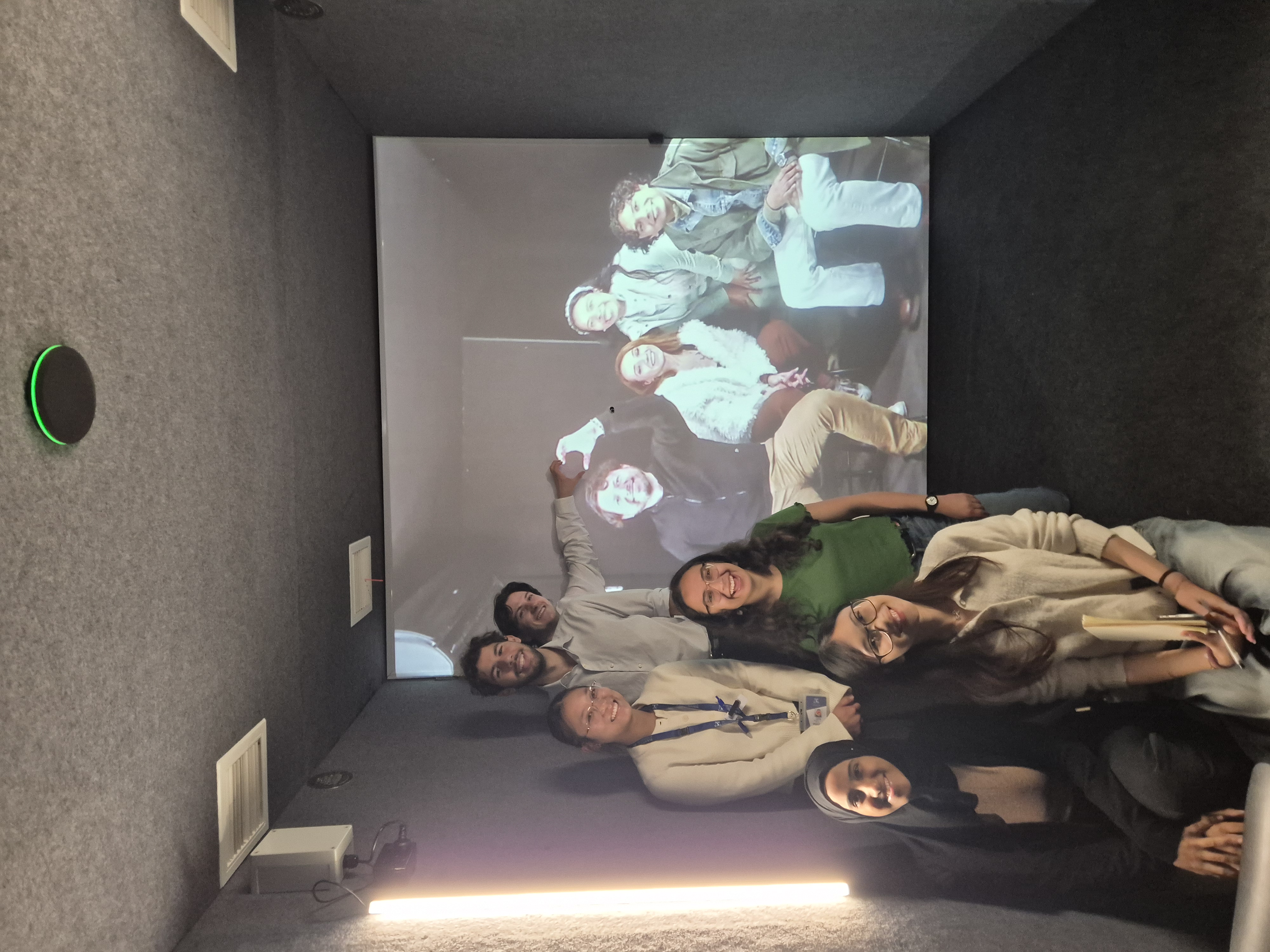 Six students stand in front of the large screen which features four people on it. One person in the room and one person on the screen match up hands in a heart shape.