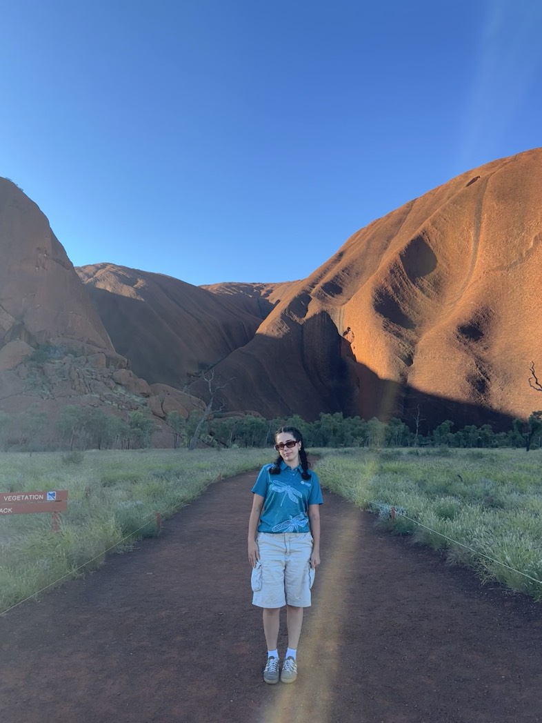 a person in a blue tshirt and beige shorts stands on a track with grass either side and orange/brown hill/rock formations behind them. The sky is bright blue.