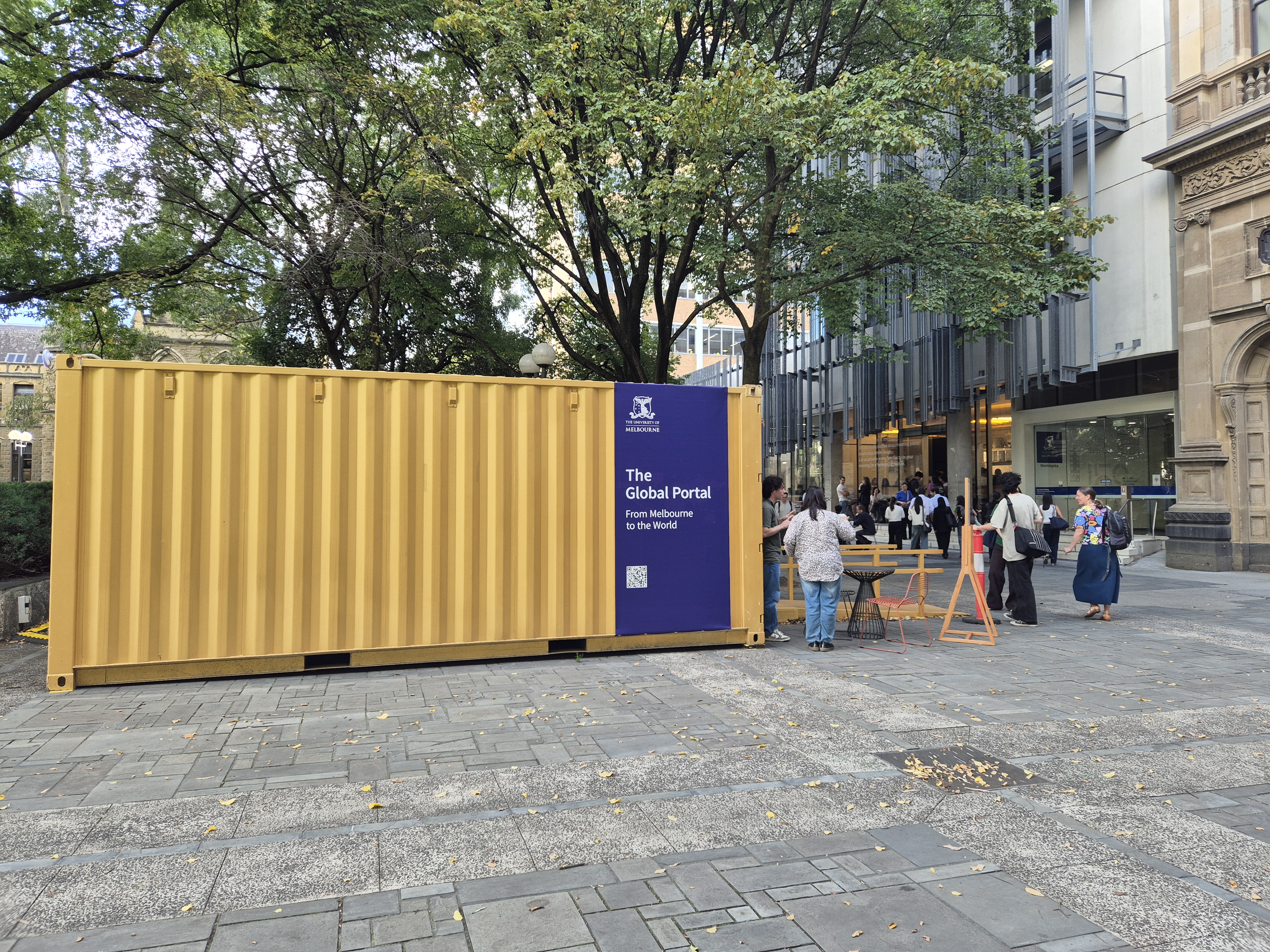 Exterior of yellow shipping container, on paved concrete ground, building in the background, trees overhead, people mingle around
