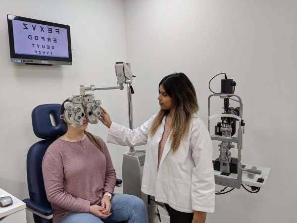 Optometrist providing an eye check at the Melbourne Eyecare Clinic, shows machinery, two people and room.