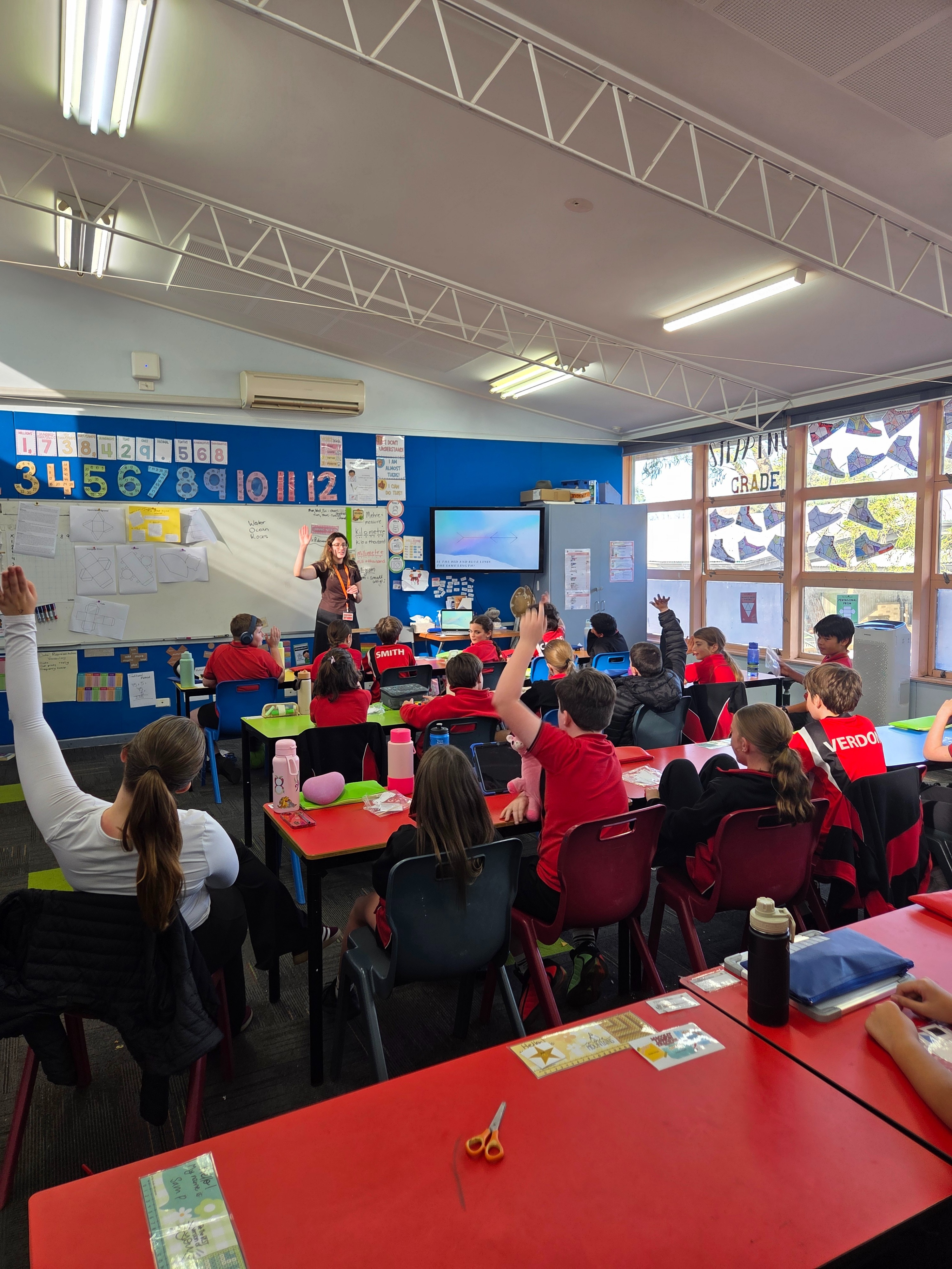classroom with children, some with hands raised, teacher at the front of the room
