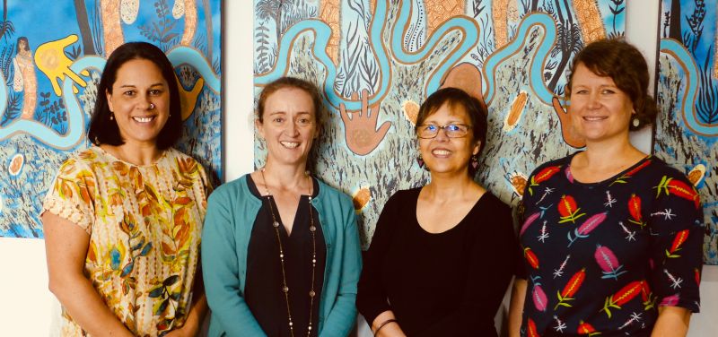 Four people standing and smiling in front of an Indigenous artwork by Treahna Hamm. From left to right, Shawana Andrews, Tamara Clements, Associate Professor Louisa Remedios and Joanne Bolton
