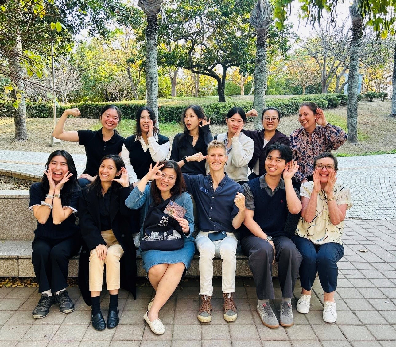 Group photo of people facing camera. Each individual is smiling and gesturing to a body part to signify their discipline (eg. ear for audiology)